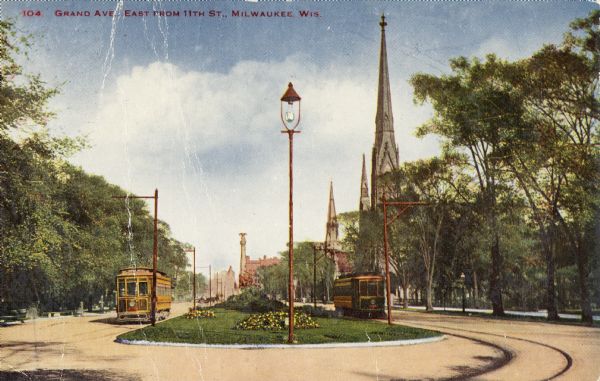 Now called Wisconsin Avenue. On the left are trees and a trolley in the road. On the right are several church towers, more trees, and another trolley. In the distance is a column of a memorial. A lamppost is located in the center foreground. Caption reads: "Grand Ave., East from 11th St., Milwaukee, Wis."