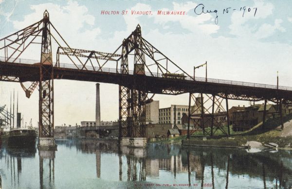 View looking up at the viaduct over the river, with a trolley on the tracks on the metal support bridge. Along the right shoreline is a grassy riverbank with industrial buildings. Caption reads: "Holton St. Viaduct, Milwaukee."