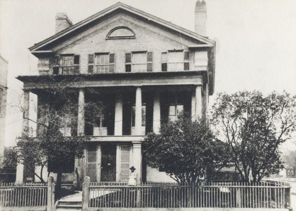 Built in 1840 by Lemuel Hull.  William Singer was the mason.  The basement was made by lowering the hill.  There were originally only two stories.   First and second levels have a columned porch.  A series of four windows line the facade of the top two floors, while the ground level has only two windows and a shuttered door.  The yard has several trees, a path from the road, and a wooden fence.  In front of the house is a small child wearing a hat.