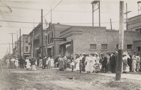 A large crowd is gathered along a sidewalk, spilling onto the road, in front of the station.