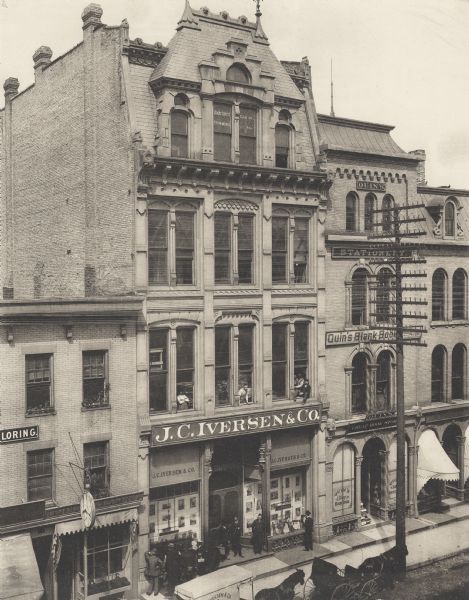 Four-level building with shorter buildings flanking it on either side.  On the road in front two carriages are visible, drawn by horses.  A group of men are in the doorway and in front of the store windows, and four other men are in the windows on the second floor.  On the right is Quin's Cheap Book Store.