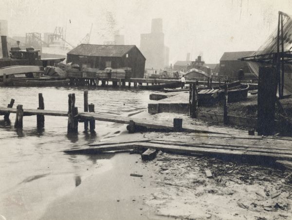 A fishing settlement. Two piers at the shoreline with a sandy beach, one with a ramp next to it.  There are several buildings in the background.  Two tugboats are next to two smaller buildings, and a man is pulling a small boat from the water.