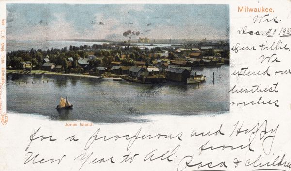 Elevated view of island with houses and boathouses and boats along the shoreline. Industrial buildings are in the background, and a sailboat is on the water in foreground. Captions read: "Milwaukee" and "Jones Island."