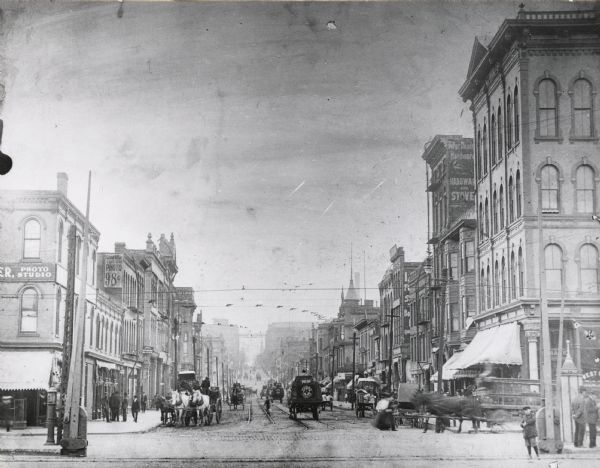 Previously known as Chestnut Street; view from the corner of 3rd Street.  Image looks down the road to the vanishing point on the horizon, where a steeple can be seen on the right.  The road is lined with buildings.  Various wires cross over the street in between the buildings, while horses, carriages, and people fill the brick and track laden road.  Signs for a photo studio are on the closest buildings on the left.