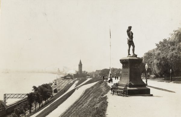 Located in Juneau Park, the statue is on the right, on the edge of a steep hill. The hillside is lined with paths, and ends next to several railroad tracks and the lake. The train station is in the middle background, and the city line is barely visible behind it on the left. The figure is on a tall pedestal, turned from the viewer, with a hand holding a horn on his hip.