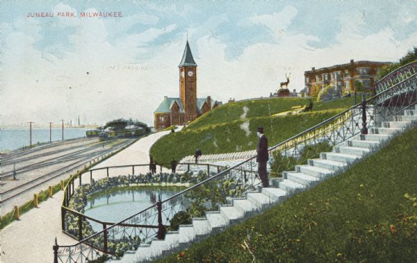 Train station in the distance, with a man standing in the foreground on steps leading down to the lake, near a fenced-in pond. On the left are railroad trains and tracks near the depot, and Lake Michigan. Caption reads: "Juneau Park, Milwaukee."