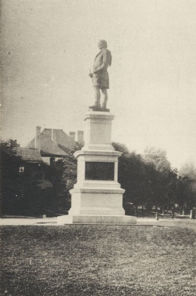 Side view of monument with plaque. Behind the statue is a road, trees, and the top of a building.  A fire hydrant and a small group of people are on the right.