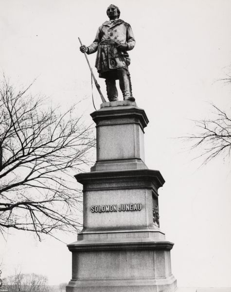 Close-up of statue in Juneau park. His right hand is holding a rifle, his left hand is resting on his belt. His name is inscribed on the middle level of the plinth.