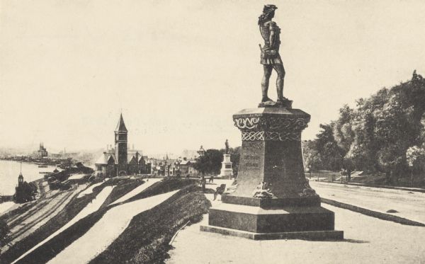 Statue of Leif Ericson is to the right of center in the foreground, with the figure facing the city.  On the left is a hill with paths, a train station, railroad tracks and trains, and Lake Michigan.