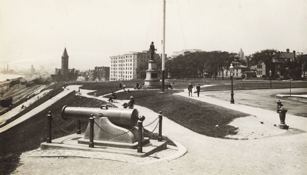 A canon is in the foreground to the left. There are a number of pedestrians in the park and on the path by the lake. In the background on the left is a train station, railroad tracks, trains, Lake Michigan, and a view of the city along the shoreline.