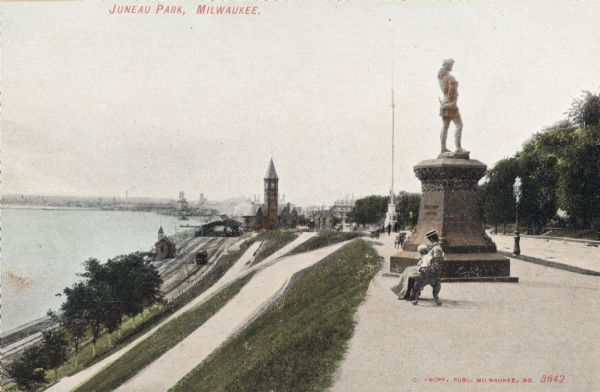 View along top of hill in the park toward a woman and child sitting on a bench near the monument. At the bottom of the hill on the left are railroad tracks, trains, and a station. The city is in the background. Caption reads: "Juneau Park, Milwaukee."