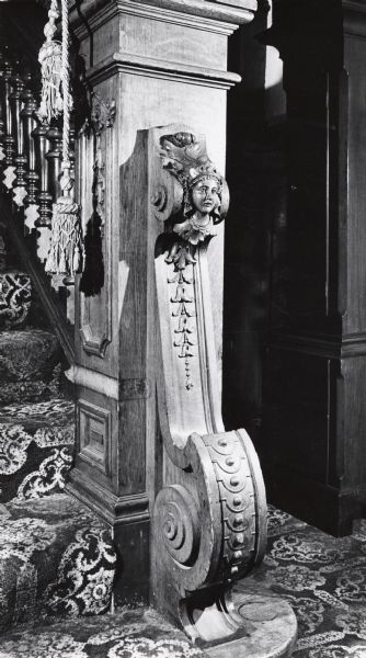 Interior of 2432 West Kilbourn Avenue. Carved wood newel post of a head with floral and curving motifs. Carpeted stairs are on the left, with large ropes and tassels hanging from the post.