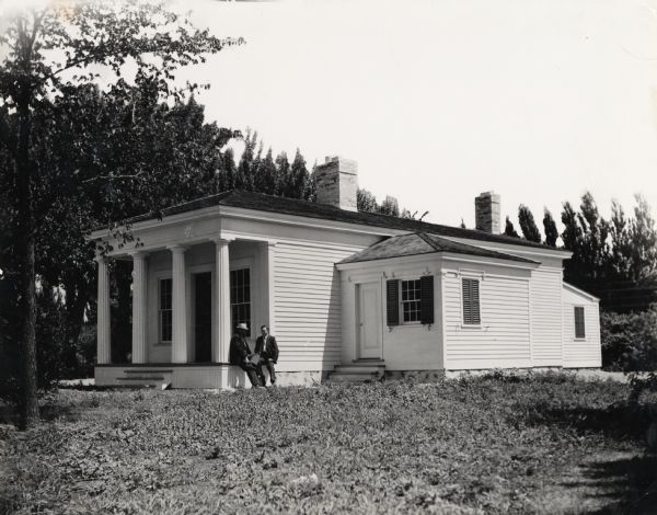Benjamin Church House.  Two men sit on the porch of a one-story white house.  Fluted columns on front porch roof, which is continuous with the main roof.  A small side room extends out from the right.