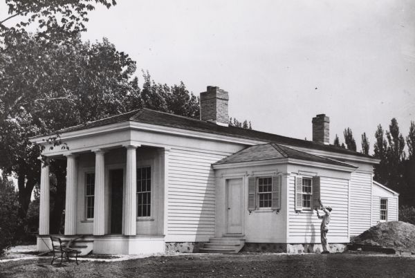 Benjamin Church House in Estabrook Park, operated as a museum by the Milwaukee County Historical Society. House is at a three-quarter angle to the viewer. Some boards are lying on the front porch stairs behind a bench, and a man is working on the shutters on the right.