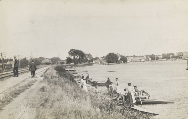Two men walk along railroad tracks near water's edge. Standing on the shoreline are a group of adults and children gathered around boats.  Buildings can be seen in the background.