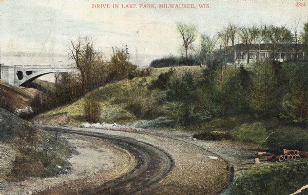 A dirt drive in the park, in between some hills, with a bridge in the background leading to a building on the right.  Next to the path are a stack of pipes, and remnants of snow. Caption reads: "Drive in Lake Park, Milwaukee, Wis."