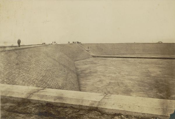 Reservoir, looking South, with pedestrians walking along the edge of the empty reservoir. A team of horses with a cart or wagon can also be seen in the background. Caption at bottom reads: "Milwaukee Water Works, Kilbourn Park Reservoir (Looking South)."