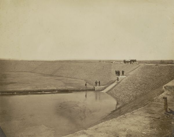 Looking down into the reservoir partially filled with water. A group of men stand at various levels; some with the carriage and horses at top, four on a ledge halfway down, two on a lower ledge, and one on a pipe above the water level. Broken barriers run along the top edge. Caption at bottom reads: "Milwaukee Water Work, Kilbourn Park Reservoir."