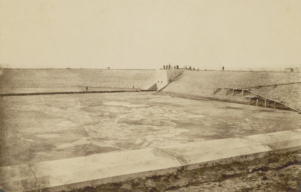 Several men are standing on top of a square ledge over the reservoir.  Other men are working in the reservoir. On the right is a wooden walkway.
