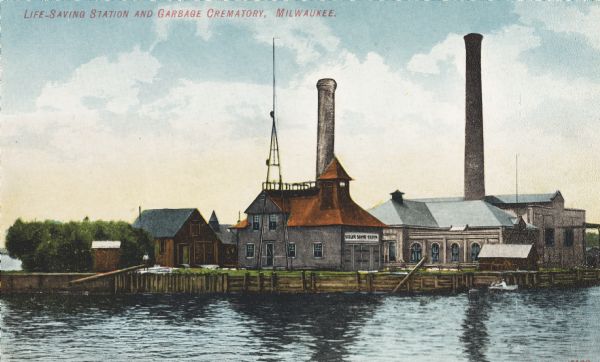 View across water toward several buildings on shoreline, two with large smokestacks. A man is standing on the shore, and two people are in a boat on the right. Caption reads: "Life-Saving Station and Garbage Crematory, Milwaukee."