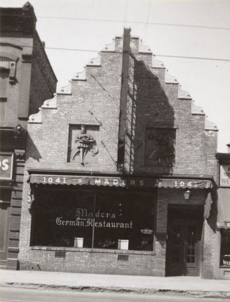 Exterior of building, with a peaked roof with embellishments on the edge. Two bas-relief figures of a man and woman carrying food and drink are set in the brick wall above the window and entrance. There is a large metal structure, presumably a sign, above the awning over the window.