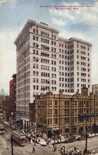 Elevated view of the hotel on a street corner. The Majestic is the tallest building in the image. Signs designate two of the buildings, with a smaller building in-between. People, cars, and trolleys line the roads and sidewalks. Caption reads: "Majestic Building and Schlitz Hotel, Milwaukee, Wis."