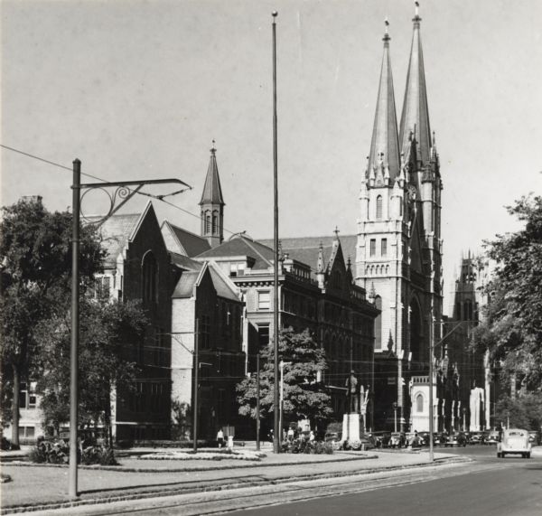 Gesu Pastoral Residence Catholic Church, 1131 W. Wisconsin Street. Church towers, with a clock, are right of center, with wings to the left and right. A monument with a statue of a man can also be seen near the parked cars.