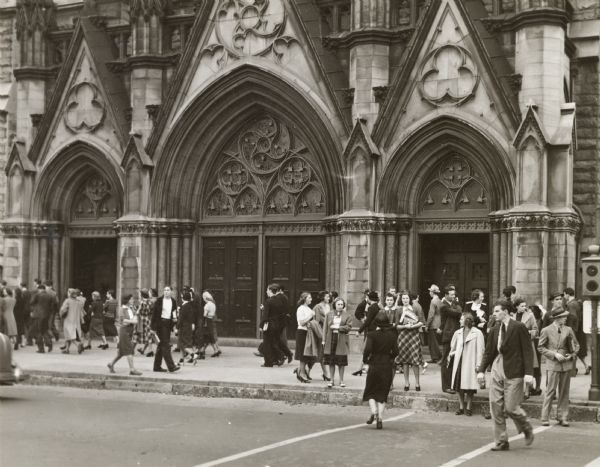 Marquette University, 1131 W. Wisconsin Street.  People are walking on the sidewalk in front of the entrance.  The three sets of doors have ornate pointed arches, and other gothic designs.