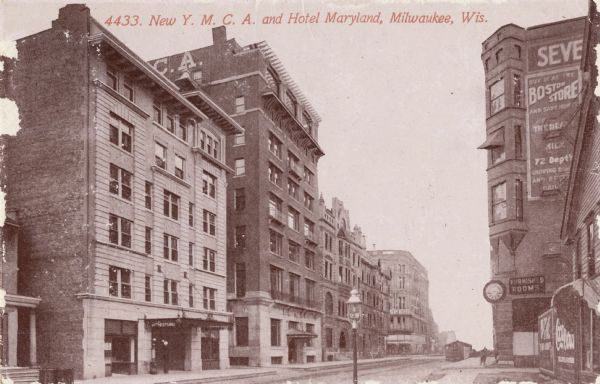 View across street toward the hotel on the left, with the Y.M.C.A. next door. On the right side of the street is a building with a billboard for the Boston Store, Coca-Cola, and furnished rooms. Caption reads: "New Y.M.C.A. and Hotel Maryland, Milwaukee, Wis."