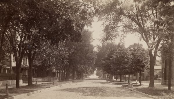 Looking down street toward Lake Michigan, which is visible at the end of the road.  The dirt street is lined with trees and houses.   On the right stands a man in a hat with a little girl on the sidewalk.