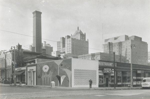 Designed by Maynard Meyer, architect. The one-story building has a mural near the entrance, and a pierced wall on the other. To the right is a restaurant, to the left is a place called Foxs. Tall buildings are in the distance.