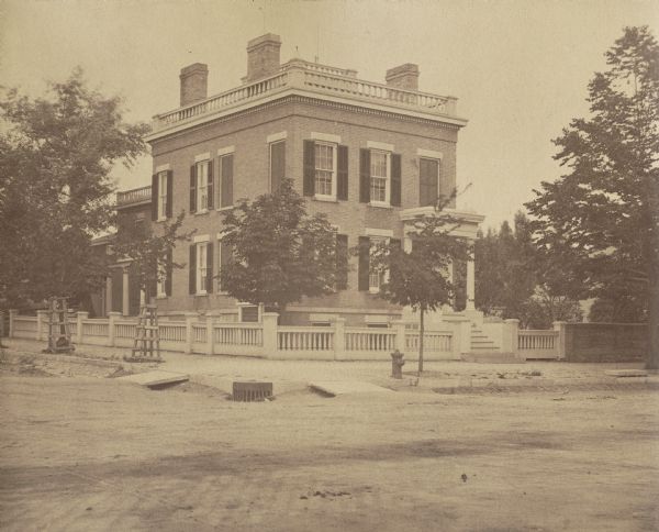 The brick home of Judge Andrew Galbraith Miller at 174 Wisconsin Street. There are steps up to the front entrance, a fence, and a fire hydrant on the sidewalk corner. The road in front is of dirt and brick.