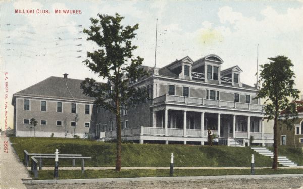 View from road toward a large building on a small hill above a road. Three posts are along the road, and small rails line the the sidewalk near a small drive. A man is standing on the grass in front of the porch. Caption reads: "Millioki Club, Milwaukee."