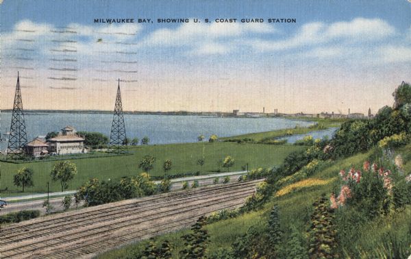 View down the hill toward the U.S. Coast Guard Station on the left with two communications towers on the Lake Michigan shoreline. Railroad tracks are just below the hill in the foreground. Caption reads: "Milwaukee Bay, Showing U.S. Coast Guard Station."