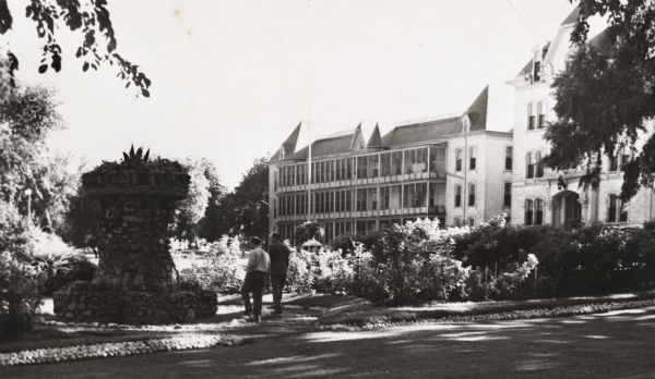 Large hospital in the background, with a large stone fountain or planter on the left. Two men are walking in the garden near a drive.
