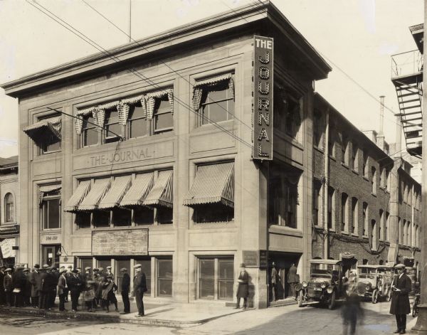The Milwaukee Journal Building located at 734 North 4th Street. To the right of the building in an alley are circulation trucks. Newsboys and customers are on the sidewalk to the left. There is a blackboard on the front of the building above windows, which has the scores of the latest baseball games, under the heading: "Read the Original Journal 'Peach.'"