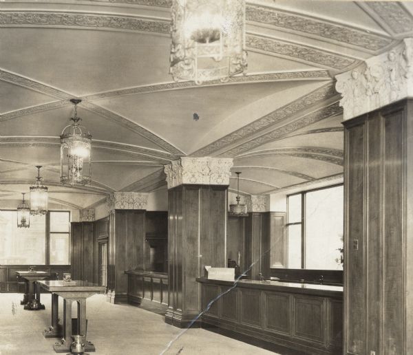 Building completed around 1926-27. Interior view of a desk area which runs along the right wall, with wood paneling on the desks and columns.  Two tall writing tables are on the left. Carved capitals top the paneled columns. Four ornate lamps are hanging from the ceiling.