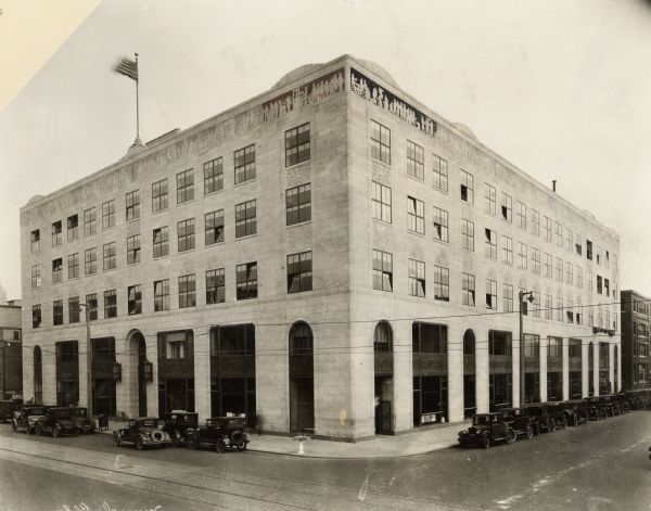 Building completed about 1926-27. Corner view with parked cars lining the street. A flag is flying from the roof. A decorative band of a classical relief lines the top of the building.