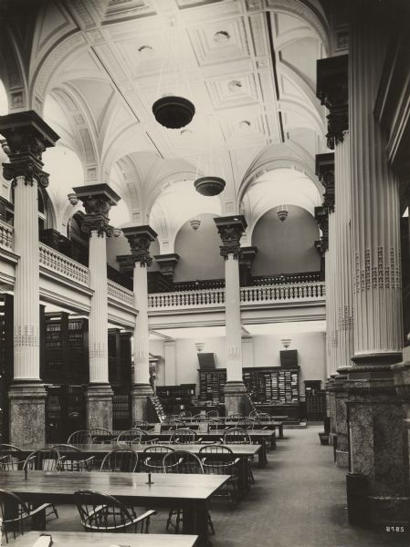 Interior of library with large open reading area and long tables and Windsor style chairs, a two-story high ceiling, and a balcony around the second level.  Fluted columns and capitals hold up the vaulted ceiling.