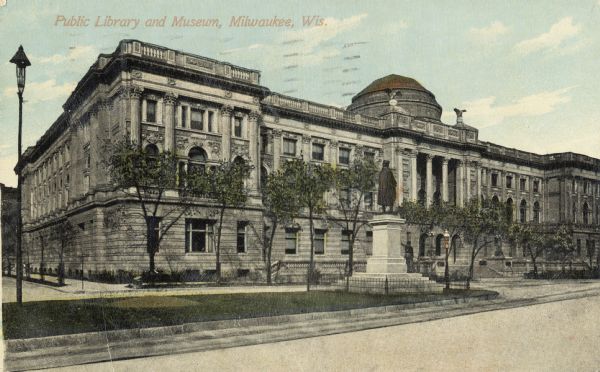 There is a dome on top of the building at the entrance. A monument is in a grassy median in the road between the building and the foreground. Caption reads: "Public Library and Museum, Milwaukee, Wis."
