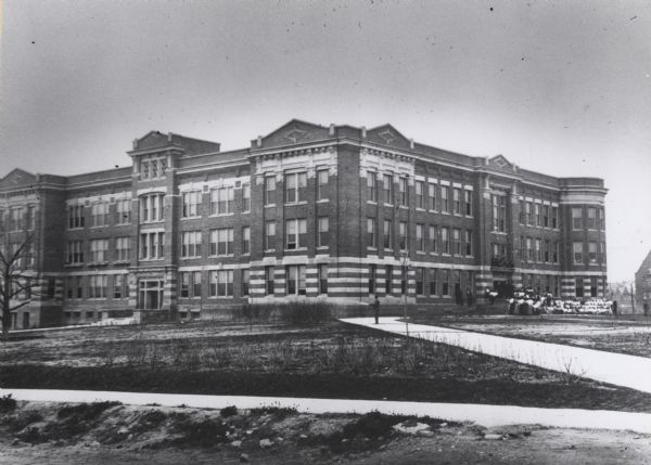 Exterior of the school with a man standing on the sidewalk in front of the three-story building spraying the lawn with a hose.  A group of people are in front of the entrance on the steps and arranged in a semi-circle.