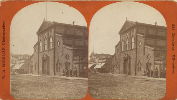 Stereograph. This building, which was destroyed by fire in 1866, was located in the commercial district known as Market Square. The Square was actually a triangular piece of land which extended from East Water Street near Mason to Market Street at Juneau Avenue.  From the middle of the nineteenth century on, it served as a social center for German-American civic life and included shops, beer halls, boardinghouses, and meeting halls. In the image, the large building has wings with roofs that step down two-levels out to each side. A group of people stand on the right.  On the left are more storefronts with awnings.
