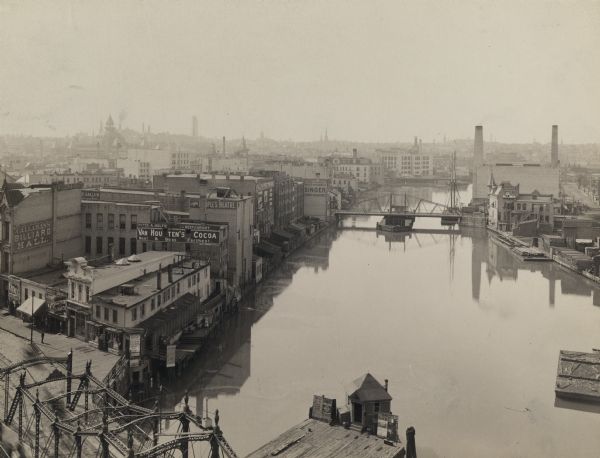 Elevated view of river looking north from the Wisconsin Avenue Bridge.  A pier with a small building and poster boards extends into the water from the bottom of the image.  Signs for Callahan's Billiard Hall, Van Houtten's Cocoa, Singer sewing machines, and a theatre are on buildings on the left side of the river.