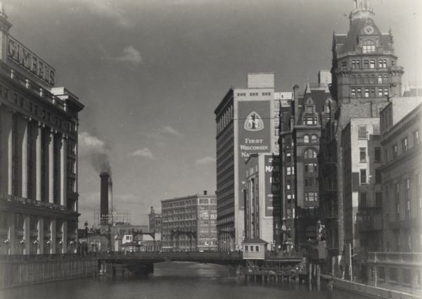 View from the Wisconsin Avenue Bridge. A building on the left has a large sign for Gimbel's on its roof. Beyond the river in the background are two large smokestacks behind a sign that says "The Electric Co". Taller buildings line the right side of the river, and one of them has a clock.