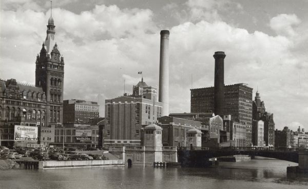 A view north of Wells Street with a bridge on the right. Two large smokestacks, and a tall building with a clock break up the skyline. On the left is a billboard for a "Coolerator." A number of cars are parked near the edge of the river on the left.