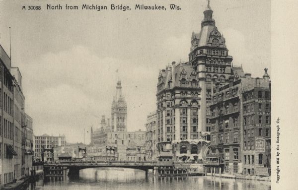 Looking north from Michigan Bridge towards another bridge. There is a clock tower on the distant right. The large building near the bridge also has a clock tower. Signs for "Hats Furs" and "J.C. Ivers..." (sign is cut off) is on the buildings lining the river. Caption reads: "North from Michigan Bridge, Milwaukee, Wis."