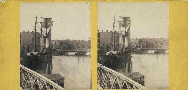 Stereograph. Part of a bridge is visible in the lower left corner. Under the bridge is a floating pier with advertisements painted on its sides. Another bridge crosses the river in the distance. A three masted sailing ship is docked next to several buildings on the left.