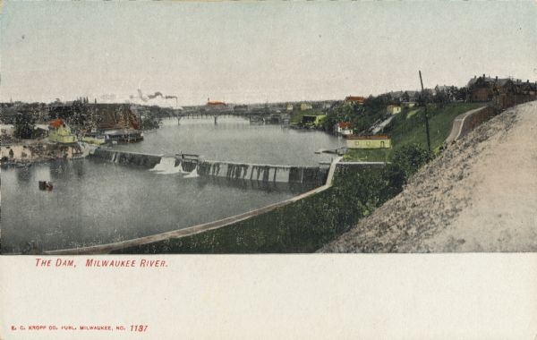 Elevated view of dam on river. A bridge is in the background. Houses are on top of the hill on the right side. Caption reads: "The Dam, Milwaukee River."