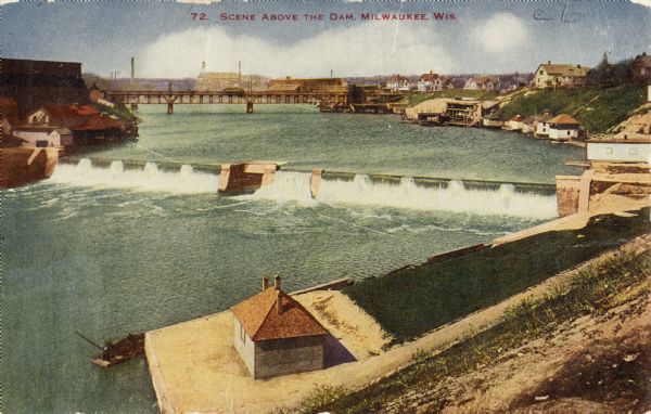 Elevated view of dam with a bridge further up the river. Buildings are along the shoreline and on top of the hill on the right. Caption reads: "Scene Above the Dam, Milwaukee, Wis."