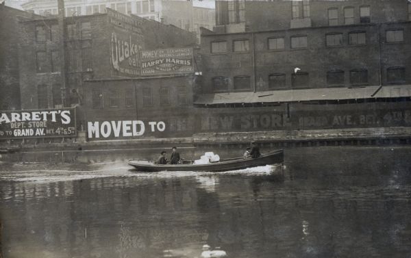 View across water of a motorboat with four men going upstream. There is a bicycle in the motorboat. The shoreline in the background has industrial buildings with advertisements for "Quaker Oats, The World's Breakfast," "Harry Harris Loan Office," and "[?]arrett's Dept. Store, Moved to the New Store, Grand Ave."